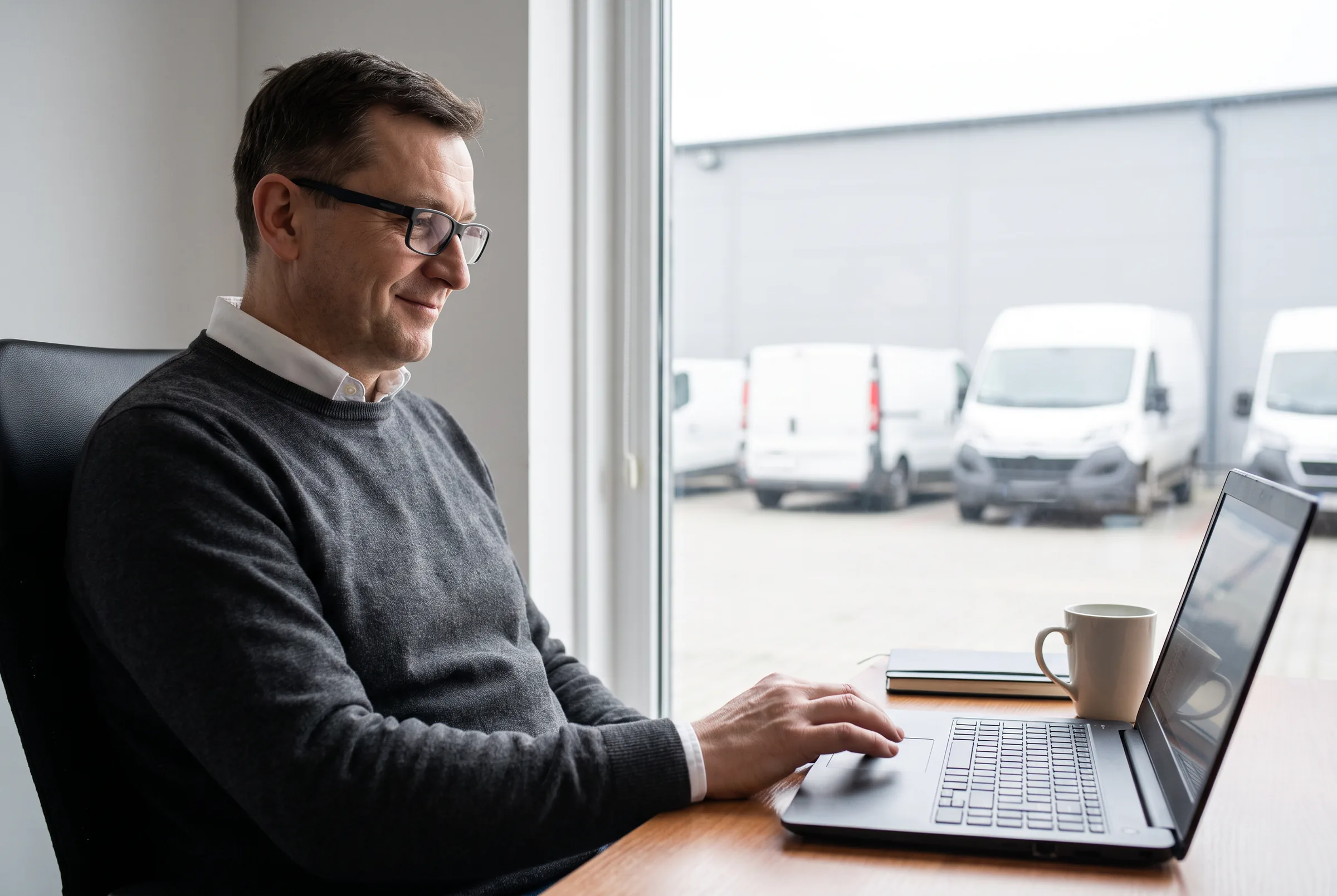 Dyrektor sprzedaży przy laptopie, za oknem dostawczaki